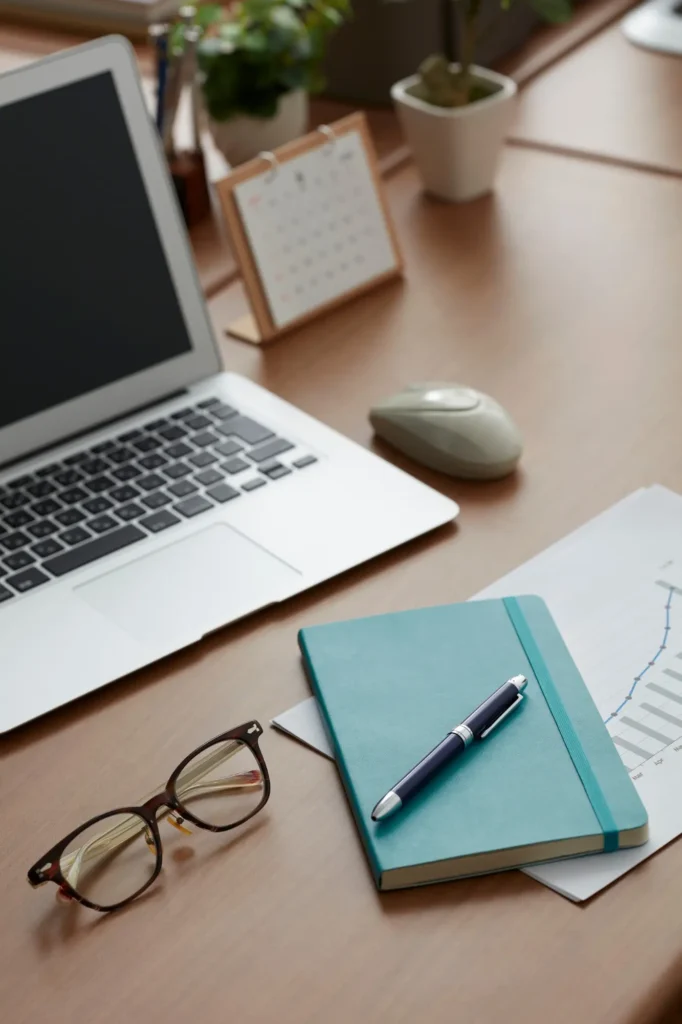 A desk with a turquoise notebook, a pen, and a laptop on top